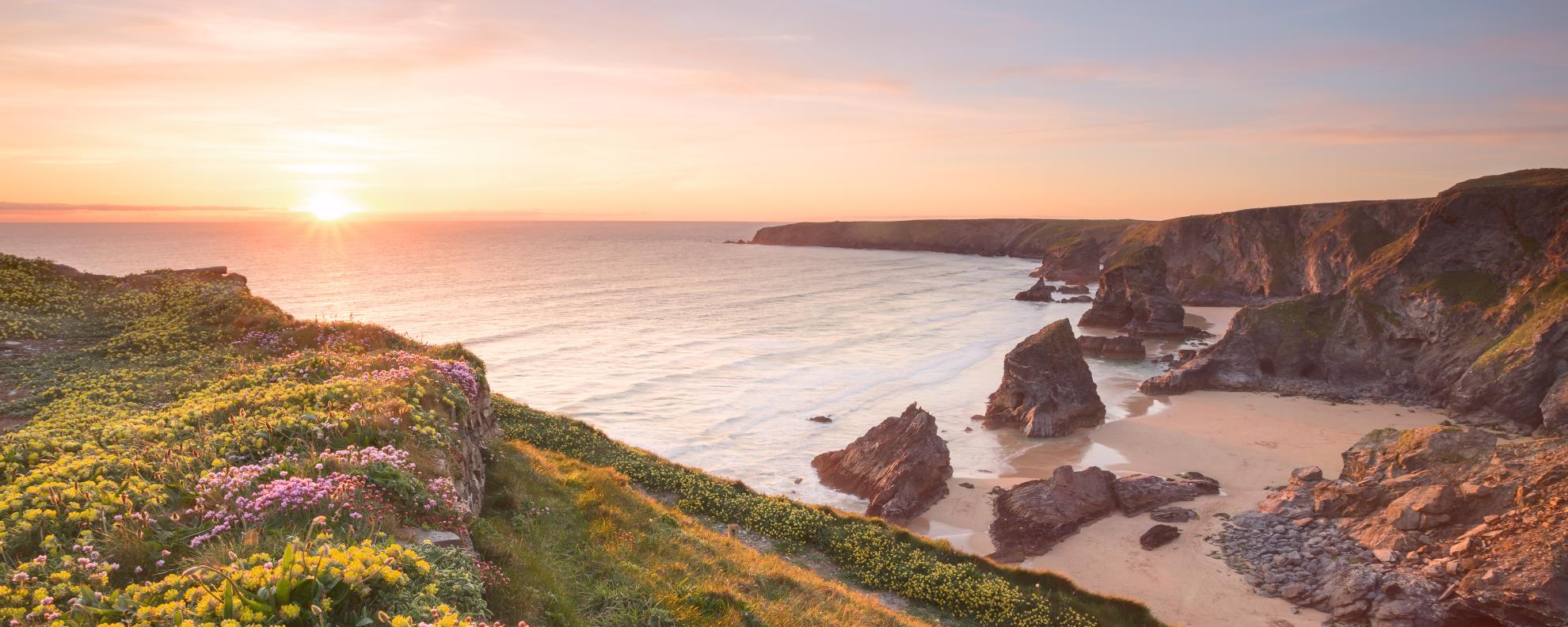 Beautiful sunset looking over Bedruthan steps in Cornwall, the sea looks tranquil and calm, the beach is empty and the green grass on the cliffs is accompanied by colourful yellow and pink flowers