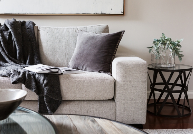 A light grey sofa in a neutrally decorated room with a dark grey blanket draped over and a book open on the sofa