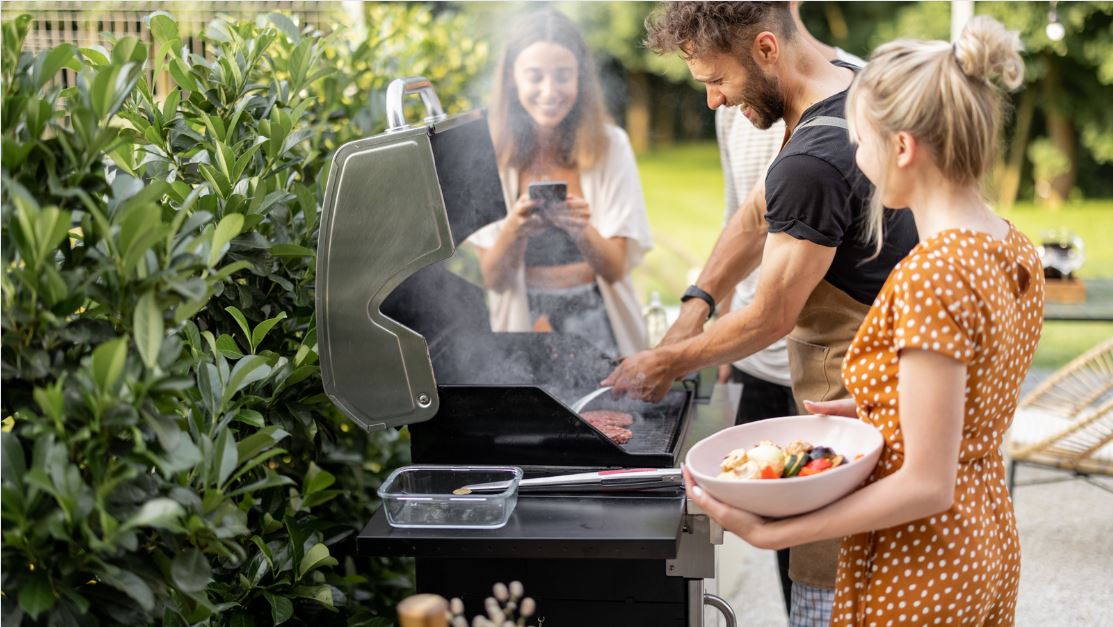 A man cooking on a BBQ in open space with two women stood either side of him smiling.