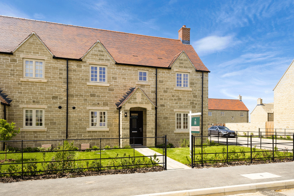 A semi-detached house, with Cotswold stone brickwork and a 'For Sale' sign outside in the front lawn