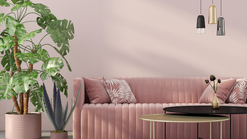 A modern living room with a pink velvet sofa adorned with patterned throw pillows, a large potted monstera plant, and an agave plant. Above the sofa hang three pendant lights in black, gold, and gray. In front of the sofa is a round coffee table with a small vase containing flowers.