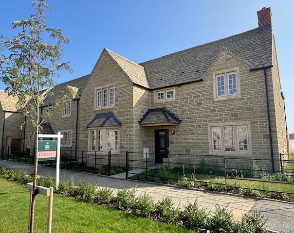 A stone two-story house with a pitched roof and multiple windows, a 'For Sale' sign on the lawn, and a tidy garden under a clear blue sky.