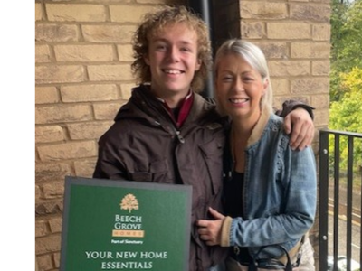 Two people standing on the balcony of their new apartment. One person is holding a green box with the text "Beech Grove Homes Your New Home Essentials".