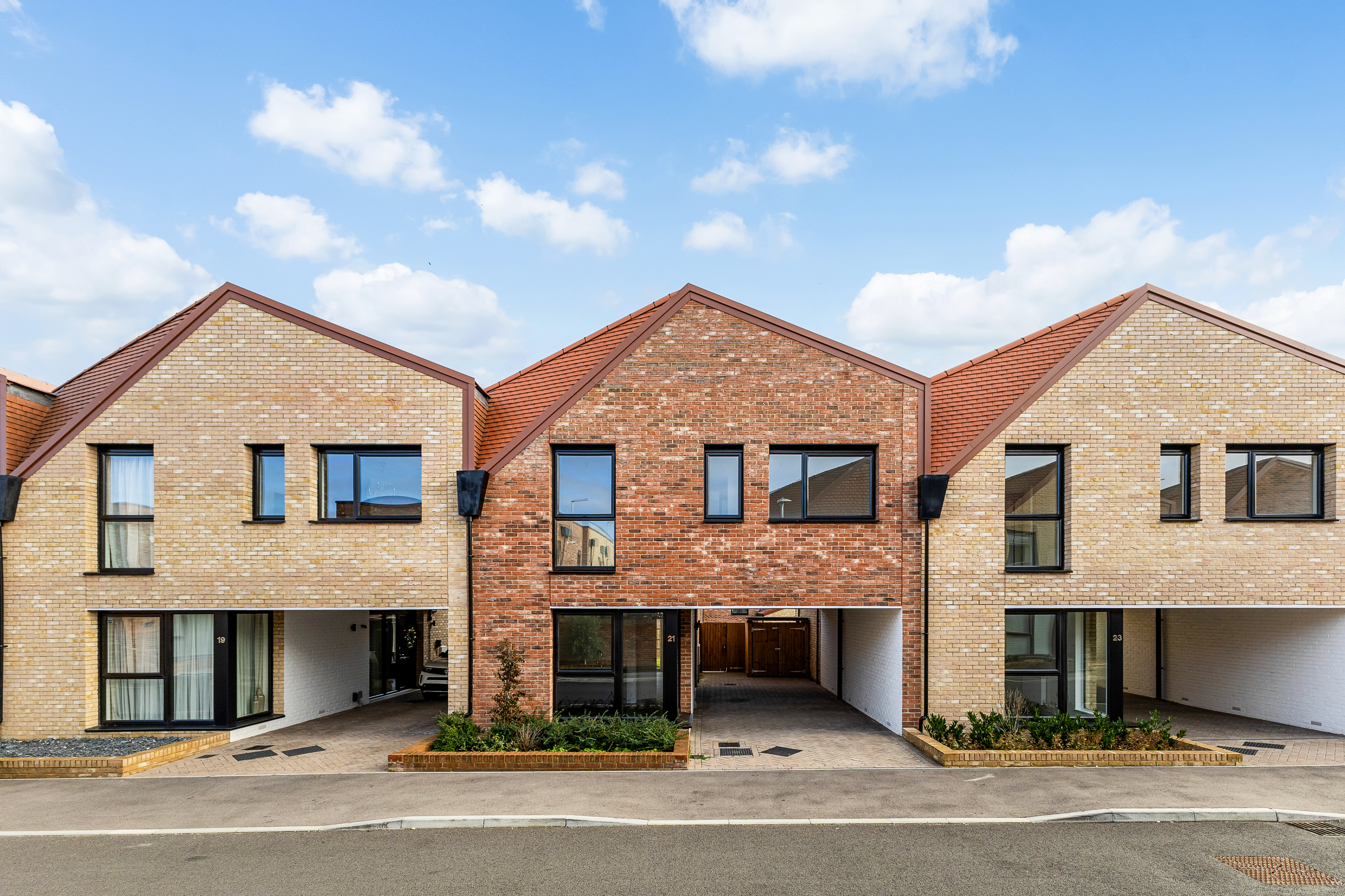 Row of modern two-storey terraced houses with light and red brick exteriors, pitched roofs, black-framed windows, and covered driveways in a new residential development under a partly cloudy sky.