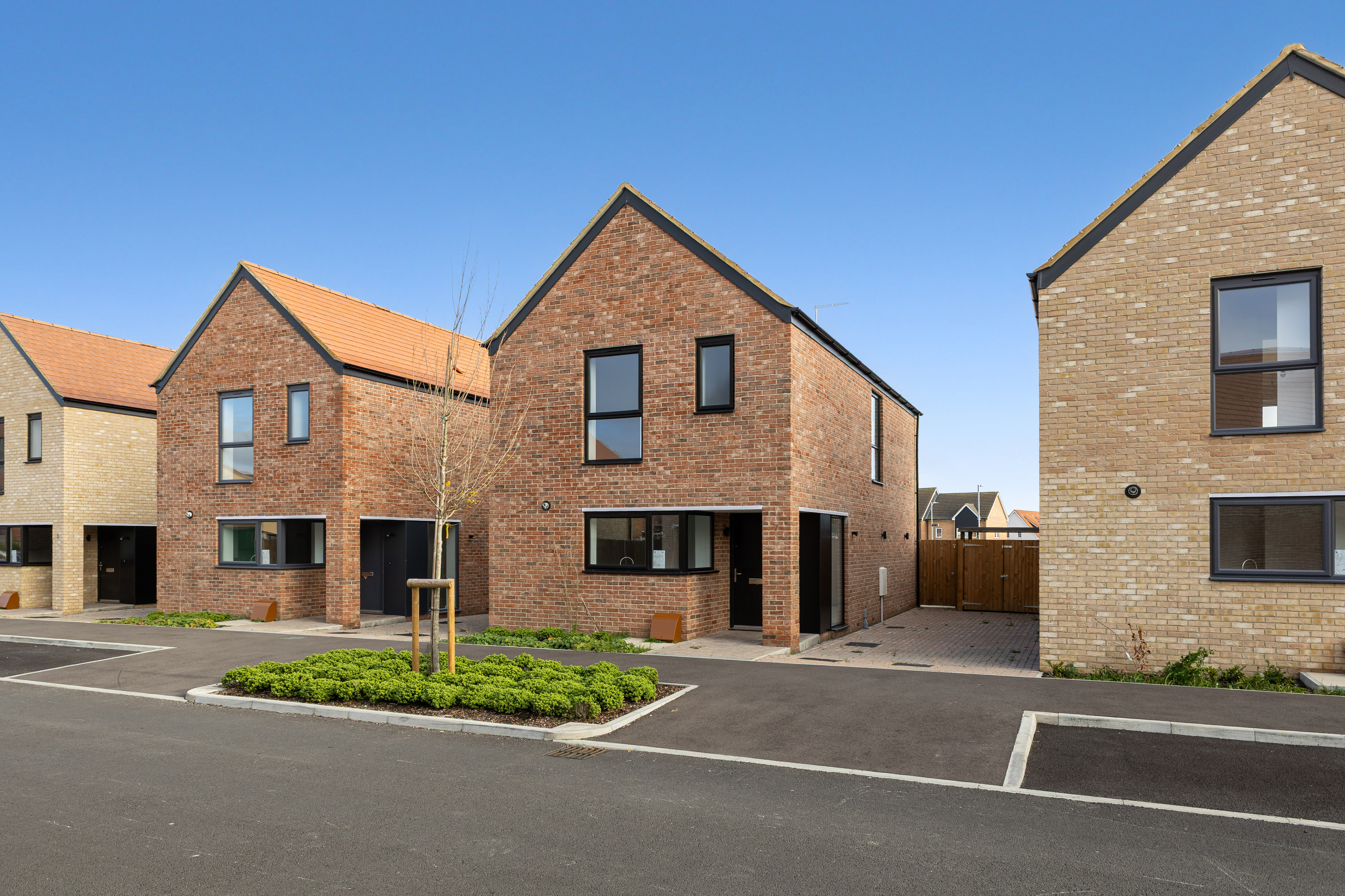 Row of modern two-storey detached houses with red and light brick exteriors, black-framed windows, and paved driveways in a new residential development under a clear blue sky.