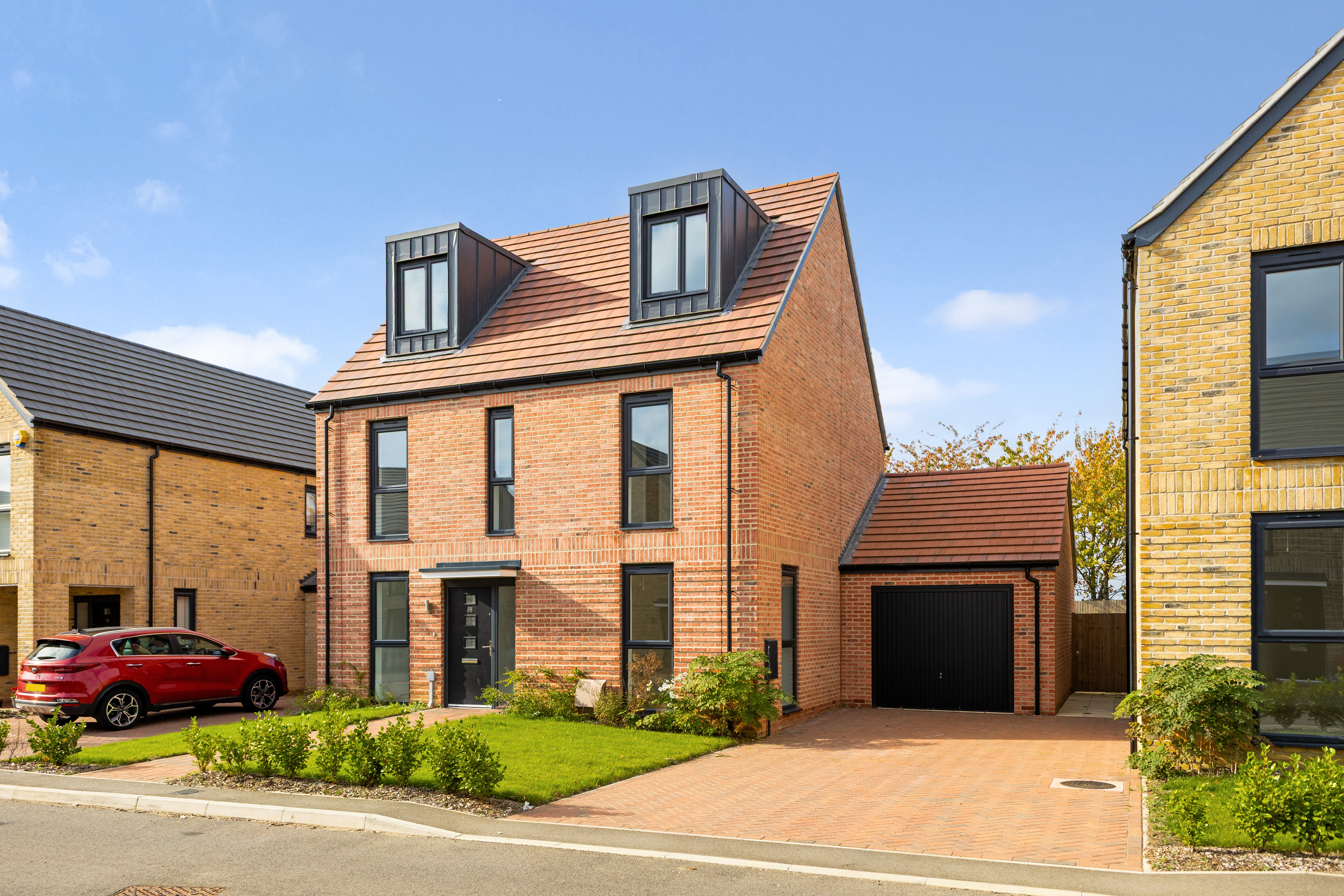 Two‑storey red brick detached house with pitched roof, front garden, driveway and separate garage, on a modern residential street.