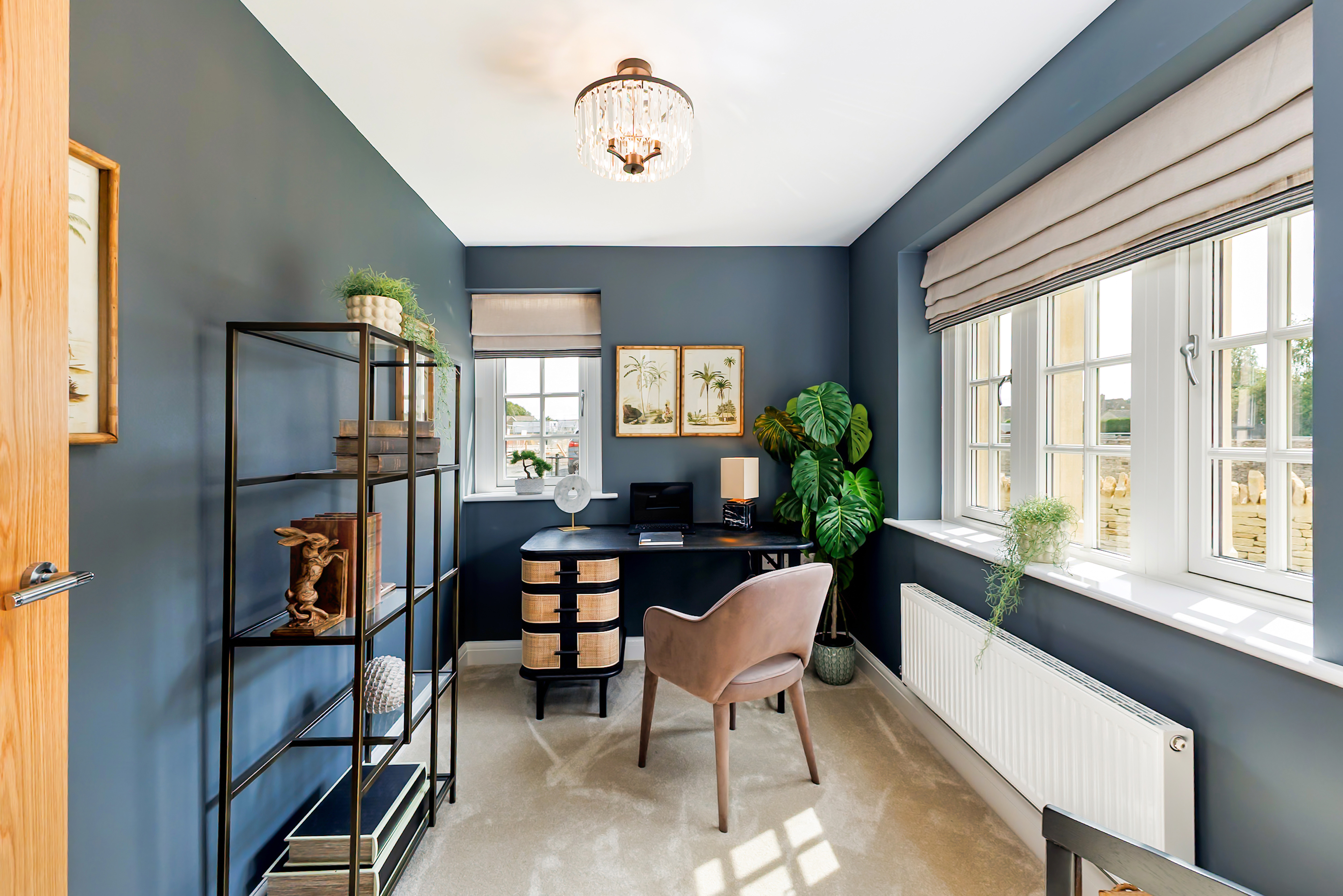 Home office in a modern new-build, featuring dark blue walls, a black desk with woven drawer fronts, a taupe upholstered chair, and a large leafy plant in the corner. A black metal shelving unit with decorative items lines one wall, while wide windows with Roman blinds let in natural light onto a soft carpeted floor, beneath a central ceiling light.