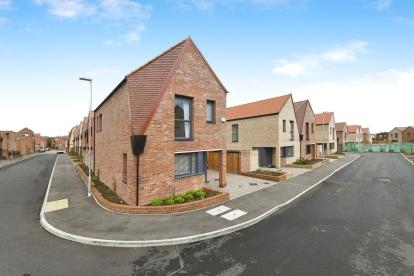 A detached, corner property with red brickwork and a red roof with dark window and door frames. The property features a carport and driveway parking and has a quirky, slanted roof