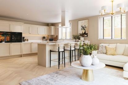 Kitchen and living space with cream cabinets, a breakfast bar with three stools, a white sofa, a round coffee table with decorative vases, and large windows allowing natural light to fill the room.