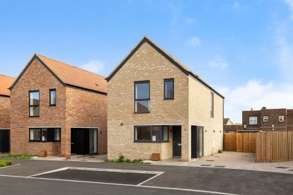 Modern two-storey detached house with light brick exterior, black-framed windows, and a paved driveway, alongside similar homes in a new residential development under a clear blue sky