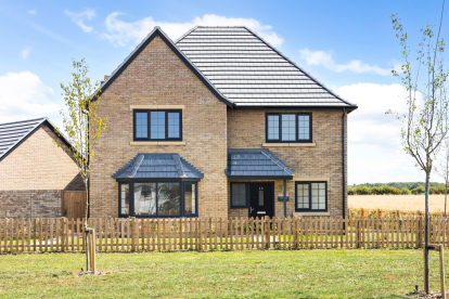 Front elevation of a modern two-storey detached new-build home with light brick exterior and a grey tiled roof. The property features black-framed windows, a central front door with canopy porch, and a bay-style window to one side. A low wooden picket fence encloses the front garden with newly planted trees and a neatly mown lawn. Open fields and hedgerows can be seen behind the property under a bright blue sky with scattered clouds.