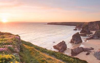 Beautiful sunset looking over Bedruthan steps in Cornwall, the sea looks tranquil and calm, the beach is empty and the green grass on the cliffs is accompanied by colorful yellow and pink flowers