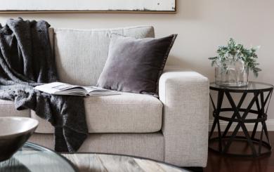 A light grey sofa in a neutrally decorated room with a dark grey blanket draped over and a book open on the sofa