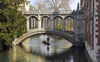 A person standing on a boat punting down the river in Cambridge about to go under a beautiful historic bridge