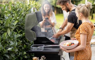 A man cooking on a BBQ in open space with two women stood either side of him smiling.