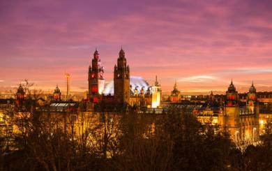 Panoramic view of Kelvingrove Art Gallery and Museum in Glasgow at sunset, with a colorful sky of purple, pink, and orange. The building is warmly lit, showcasing its towers and domes, with trees in the foreground