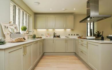 Modern kitchen with light beige cabinets, white marble countertops, and stainless steel appliances. The space features a wooden floor, recessed ceiling lights, and a central island with utensils. A large clock hangs on the right wall.