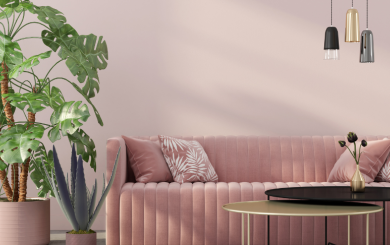 A modern living room with a pink velvet sofa adorned with patterned throw pillows, a large potted monstera plant, and an agave plant. Above the sofa hang three pendant lights in black, gold, and gray. In front of the sofa is a round coffee table with a small vase containing flowers.