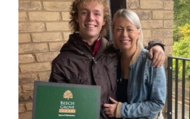 Two people standing on the balcony of their new apartment. One person is holding a green box with the text "Beech Grove Homes Your New Home Essentials".