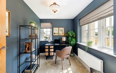 Home office in a modern new-build, featuring dark blue walls, a black desk with woven drawer fronts, a taupe upholstered chair, and a large leafy plant in the corner. A black metal shelving unit with decorative items lines one wall, while wide windows with Roman blinds let in natural light onto a soft carpeted floor, beneath a central ceiling light.
