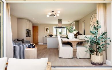 Open-plan kitchen and dining area featuring a rustic vintage-style wooden dining table with slipcovered chairs, set against a neutral-toned interior with cabinetry and striped armchairs.