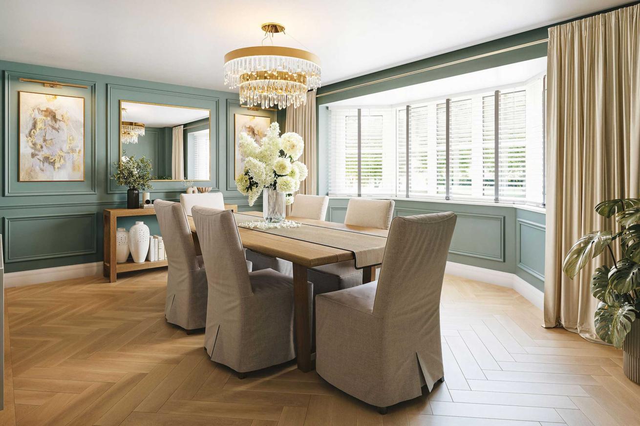Elegant dining room with a rectangular wooden table surrounded by six upholstered beige chairs, a crystal chandelier overhead, large bay window with white blinds, green panelled walls, and decorative artwork and vases.