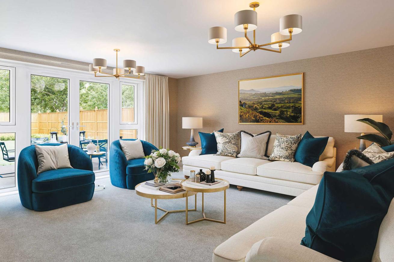 Spacious living room with large glass doors opening to a garden, featuring a white sofa with patterned cushions, two deep blue armchairs, and a pair of round marble coffee tables with gold frames. The room has neutral walls, a landscape painting above the sofa, modern ceiling lights with multiple shades, and light gray carpet flooring.