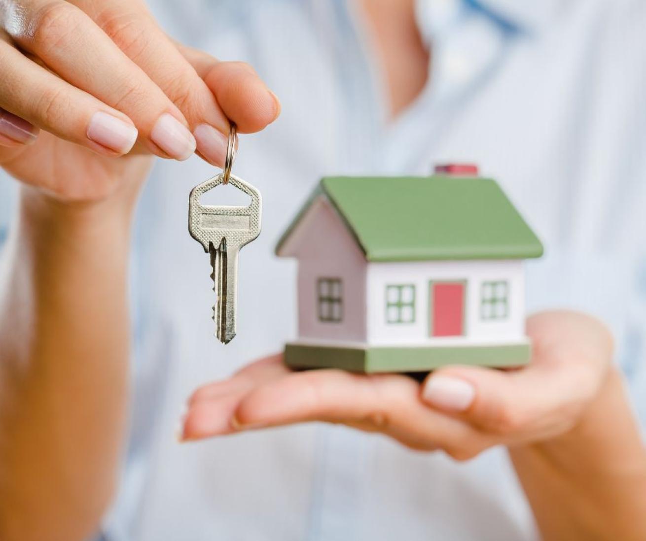 A woman holding a house key in her right hand next to a miniature ceramic house in the palm of her left hand.