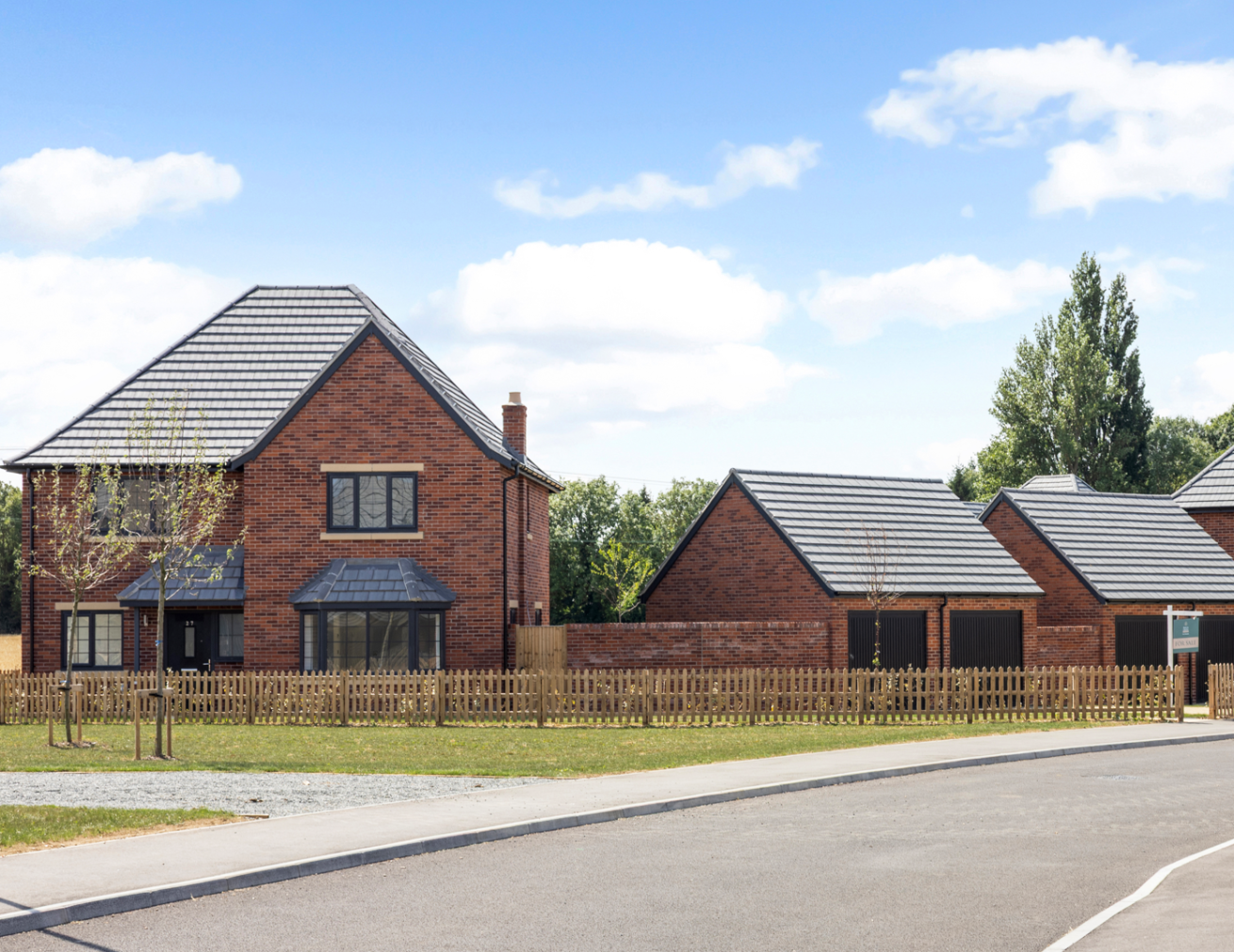 Red brick detached house with grey tiled roof, front garden and picket fence, set along a quiet street with matching garages nearby.