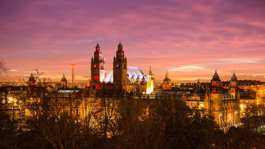 Panoramic view of Kelvingrove Art Gallery and Museum in Glasgow at sunset, with a colorful sky of purple, pink, and orange. The building is warmly lit, showcasing its towers and domes, with trees in the foreground
