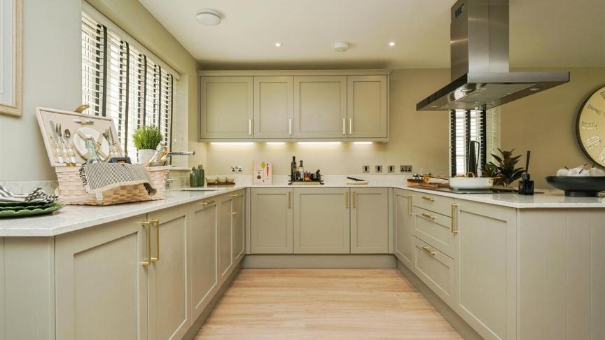 Modern kitchen with light beige cabinets, white marble countertops, and stainless steel appliances. The space features a wooden floor, recessed ceiling lights, and a central island with utensils. A large clock hangs on the right wall.