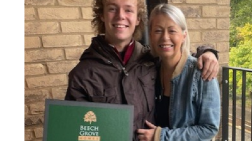Two people standing on the balcony of their new apartment. One person is holding a green box with the text "Beech Grove Homes Your New Home Essentials".