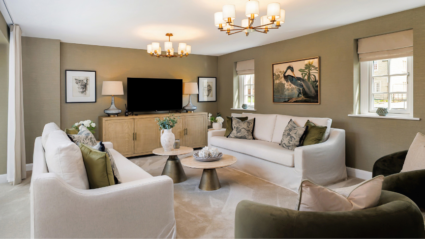 A stylish living room with two white sofas and two green armchairs arranged around a circular coffee table. The room has beige walls, three windows with Roman shades, and a large flat-screen TV mounted above a wooden cabinet. Two framed artworks and table lamps sit on either side of the TV. Two elegant chandeliers hang from the ceiling, enhancing the room’s sophisticated feel.