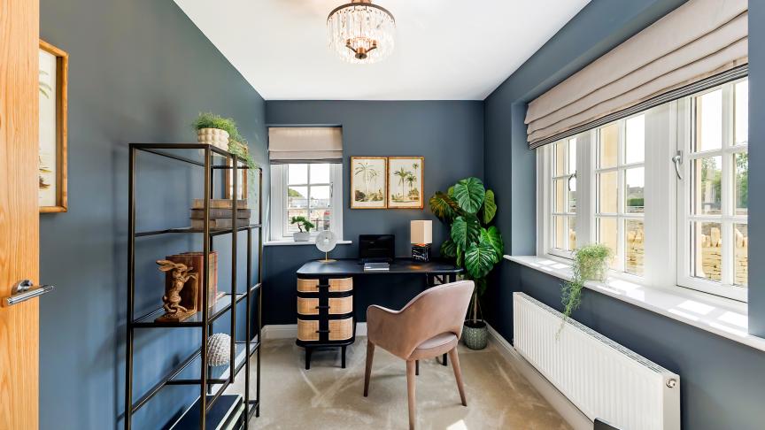 Home office in a modern new-build, featuring dark blue walls, a black desk with woven drawer fronts, a taupe upholstered chair, and a large leafy plant in the corner. A black metal shelving unit with decorative items lines one wall, while wide windows with Roman blinds let in natural light onto a soft carpeted floor, beneath a central ceiling light.