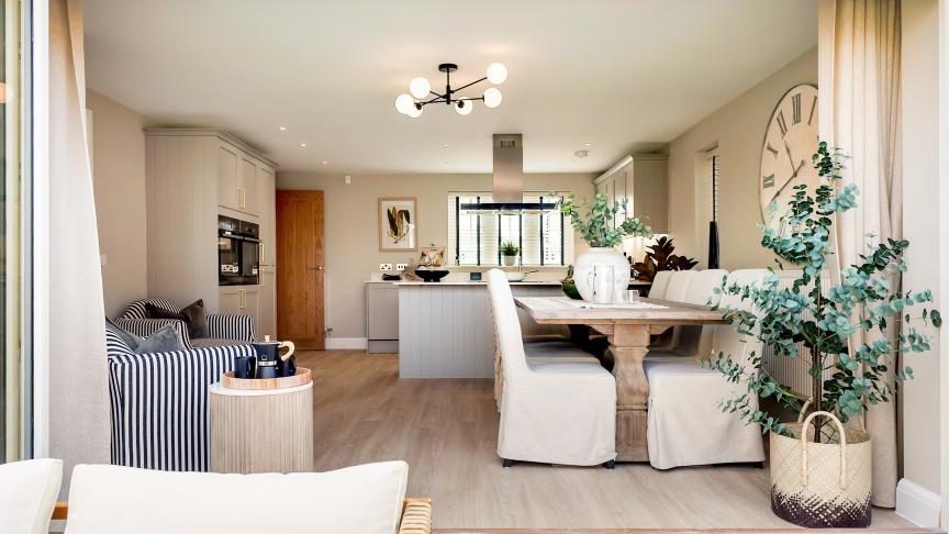 Open-plan kitchen and dining area featuring a rustic vintage-style wooden dining table with slipcovered chairs, set against a neutral-toned interior with cabinetry and striped armchairs.