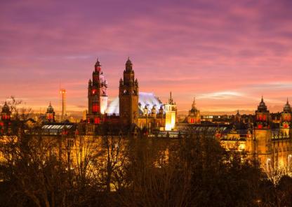 Panoramic view of Kelvingrove Art Gallery and Museum in Glasgow at sunset, with a colorful sky of purple, pink, and orange. The building is warmly lit, showcasing its towers and domes, with trees in the foreground