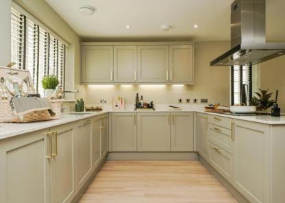 Modern kitchen with light beige cabinets, white marble countertops, and stainless steel appliances. The space features a wooden floor, recessed ceiling lights, and a central island with utensils. A large clock hangs on the right wall.