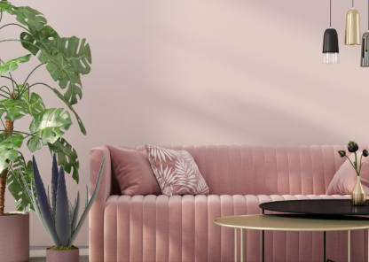 A modern living room with a pink velvet sofa adorned with patterned throw pillows, a large potted monstera plant, and an agave plant. Above the sofa hang three pendant lights in black, gold, and gray. In front of the sofa is a round coffee table with a small vase containing flowers.