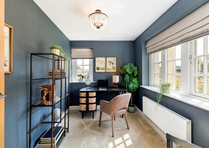 Home office in a modern new-build, featuring dark blue walls, a black desk with woven drawer fronts, a taupe upholstered chair, and a large leafy plant in the corner. A black metal shelving unit with decorative items lines one wall, while wide windows with Roman blinds let in natural light onto a soft carpeted floor, beneath a central ceiling light.