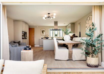 Open-plan kitchen and dining area featuring a rustic vintage-style wooden dining table with slipcovered chairs, set against a neutral-toned interior with cabinetry and striped armchairs.